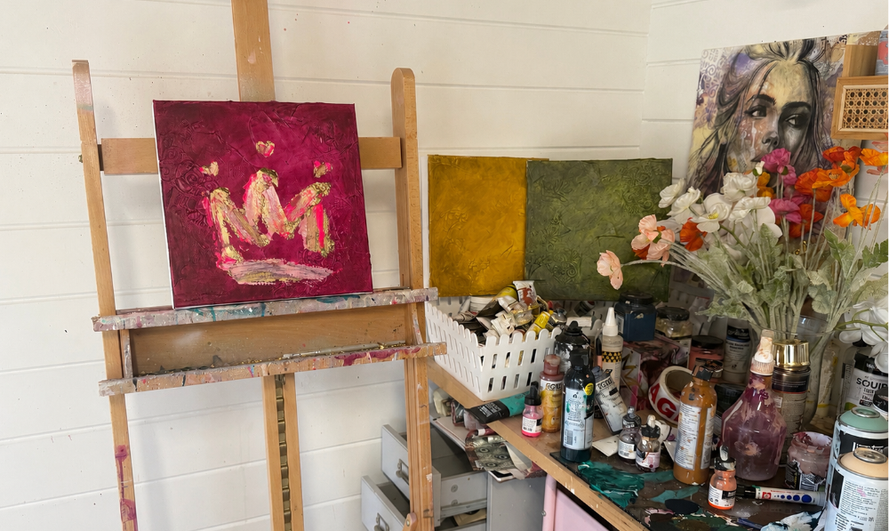 An art studio corner with a wooden easel holding a red painting of two figures, a cluttered artist's table with paint bottles, brushes, flowers, and canvases against a white paneled wall.
